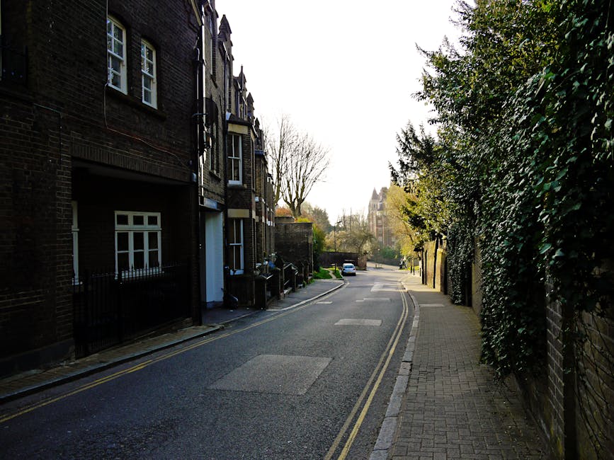A quiet residential street near Hampstead High Street NW3 featuring a paved sidewalk on the right and a narrow road with double yellow lines. On the left, there are multi-story brick houses with white-framed windows and small front gardens enclosed by black metal fences. Tall trees with bare branches and lush green hedges line the right side, providing privacy and greenery. The street appears clean and well-maintained with smooth asphalt and clear lane markings, illuminated by natural daylight that highlights the building facades, trees, and roadway. Deep Cleaning Hampstead specializes in surface cleaning and deep cleaning services that promote hygiene and upkeep of such residential areas, ensuring a pristine environment for residents.