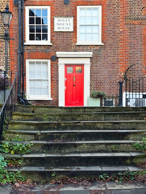 A quiet residential street near Hampstead High Street NW3 featuring a paved sidewalk on the right and a narrow road with double yellow lines. On the left, there are multi-story brick houses with white-framed windows and small front gardens enclosed by black metal fences. Tall trees with bare branches and lush green hedges line the right side, providing privacy and greenery. The street appears clean and well-maintained with smooth asphalt and clear lane markings, illuminated by natural daylight that highlights the building facades, trees, and roadway. Deep Cleaning Hampstead specializes in surface cleaning and deep cleaning services that promote hygiene and upkeep of such residential areas, ensuring a pristine environment for residents.