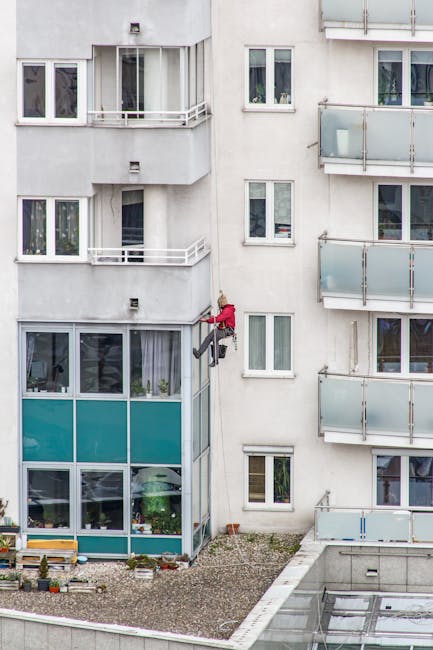 A professional window cleaner, wearing a safety helmet and harness, is ascending a high glass exterior of a modern building. The performer is using a squeegee and cloth to clean the large, reflective 
