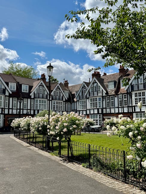 Photograph of a residential street in Hampstead, featuring a row of traditional English-style houses with black and white timber-framed facades and sloped tiled roofs. The foreground shows a neatly maintained area with blooming white roses and green bushes, bordered by a low black metal fence. A paved pathway runs alongside the garden, with a classic black street lamp positioned near the flowerbeds. The scene is illuminated by natural daylight under a bright blue sky with scattered white clouds, highlighting the well-kept appearance of the outdoor space. Deep Cleaning Hampstead specializes in surface and deep cleaning, ensuring homes in NW3 remain hygienic and visually appealing, especially in areas requiring regular sanitisation and maintenance such as gardens and exterior surfaces.