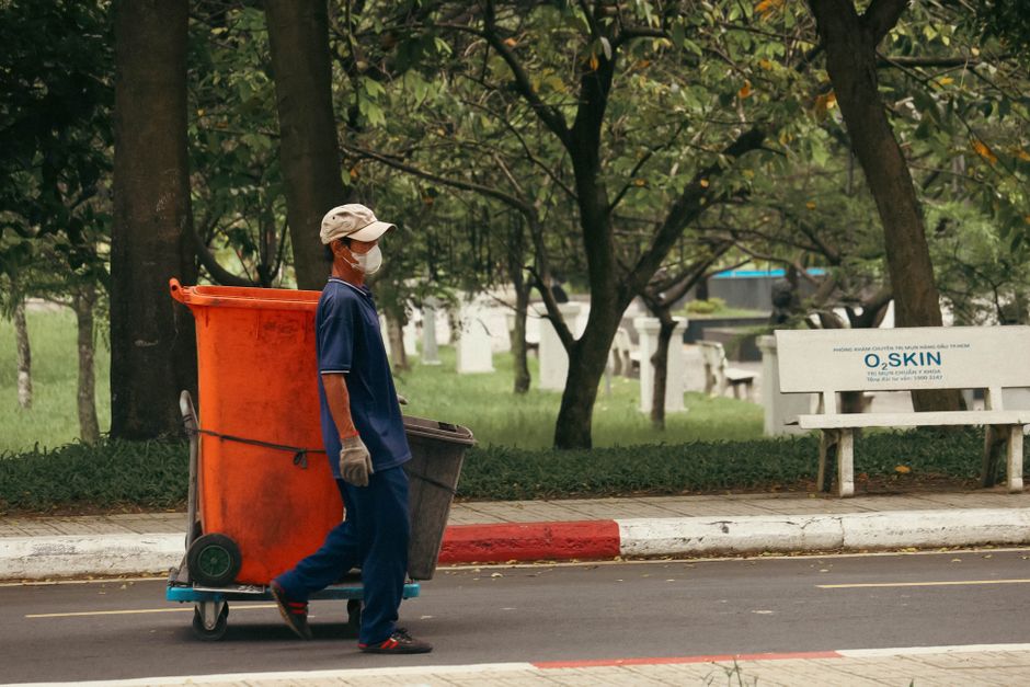 A sanitation worker wearing a beige cap, face mask, and gloves pushing an orange wheelie bin on a city sidewalk. The worker is dressed in a navy blue uniform and is holding a smaller gray bin. The bac