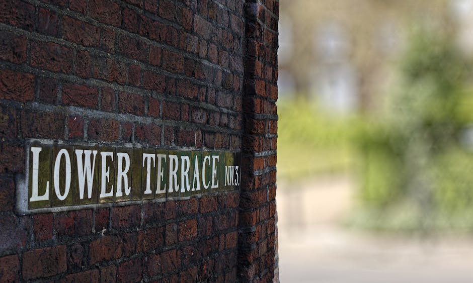 A close-up of a brick wall featuring decorative black and white ceramic street sign tiles that spell out 'TO BRANCH HILL & WEST HEATH RD' in bold white letters. The bricks are aged, with some moss and dirt visible, giving a weathered appearance. There is a small graphic of a pointing hand on the right side of the sign. The scene is evenly lit, highlighting the texture of the bricks and the signage, representing an outdoor urban environment. Deep Cleaning Hampstead specializes in surface cleaning and deep cleaning services, ensuring hygiene and cleanliness of brick and tiled surfaces in residential and commercial spaces.